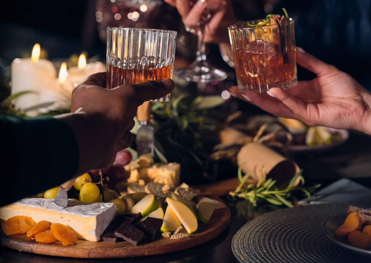 Two people cheer with a glass of whiskey, a whiskey cocktail and plates with snack on the table